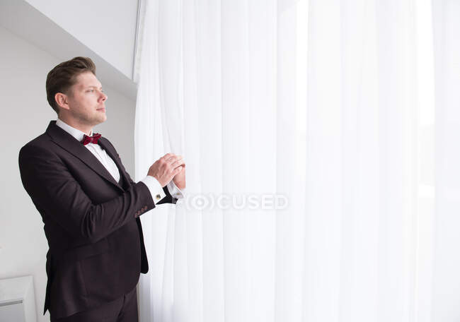 Vue latérale de jeune bel homme debout dans la chambre en costume formel noir et regardant par la fenêtre derrière le rideau blanc — Photo de stock