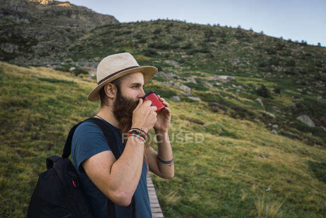 Jovem de chapéu em pé nas montanhas com copo — Fotografia de Stock