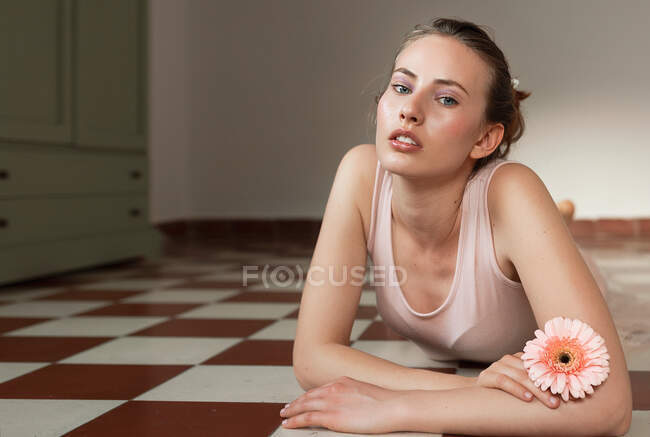 Красива молода жінка, перетинаючи руки, тримає квітку, розміщуючи на кухні плиткою підлогу і дивлячись на камеру — стокове фото