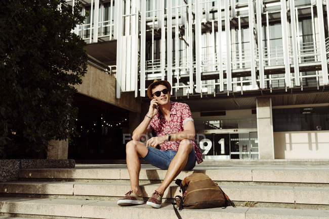 Молодой турист разговаривает на смартфоне и сидит на ступеньках в городе — стоковое фото