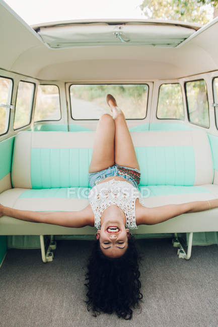 Cheerful young brunette woman lying on back seat of retro van during trip in nature — Stock Photo
