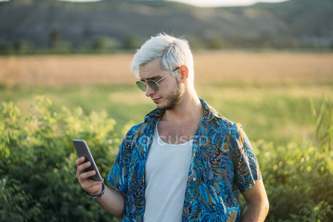 Beau jeune homme en tenue élégante riant et naviguant smartphone tout en se tenant sur fond de belle nature le jour ensoleillé — Photo de stock