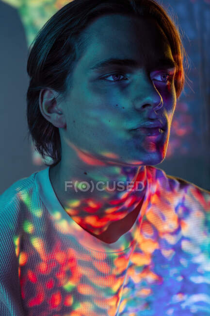 Young androgynous guy illuminated with colorful sports of bright light looking away while standing on black background — Stock Photo