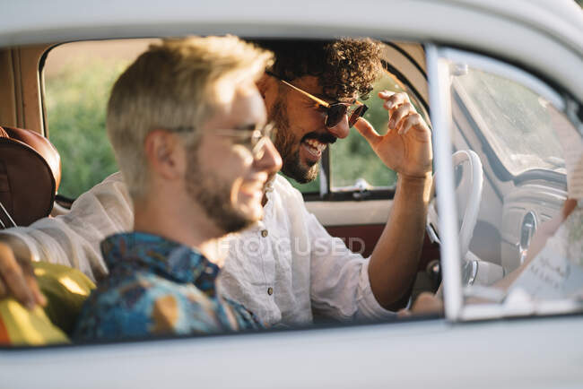 Dos chicos jóvenes riendo y sosteniendo el mapa mientras están sentados dentro de un coche retro durante un buen viaje - foto de stock