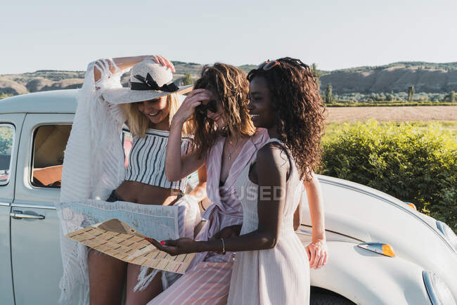 Mujeres multiétnicas de moda de pie cerca de coche vintage y mapa de lectura mientras viajan juntos en verano - foto de stock