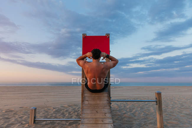 Vue arrière du gars musclé torse nu qui fait des craquements abdominaux sur une glissière en bois sur la plage au coucher du soleil — Photo de stock