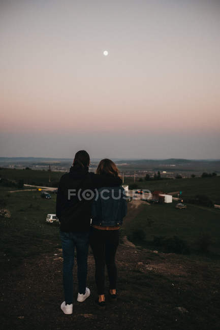 Rückansicht eines jungen Mannes und einer jungen Frau, die sich umarmen und Autos betrachten, die das Tal überqueren, während sie abends auf einem Hügel stehen — Stockfoto