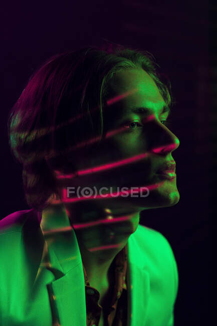 Attractive androgynous man keeping eyes closed while standing under colorful illumination in dark room — Stock Photo