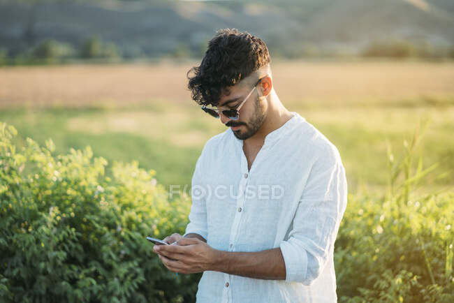 Bonito cara jovem em roupa elegante rindo e navegando smartphone enquanto em pé no fundo da bela natureza no dia ensolarado — Fotografia de Stock