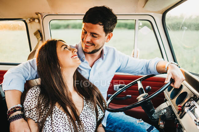 Cheerful young man and woman embracing and riding in vintage van on road in rural environment — Stock Photo