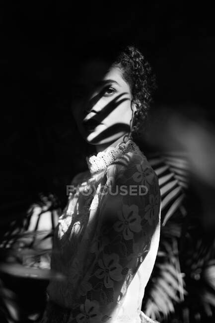 Plan noir et blanc de charmante jeune femme afro-américaine en tissus clairs avec ombre sur le visage regardant la caméra — Photo de stock
