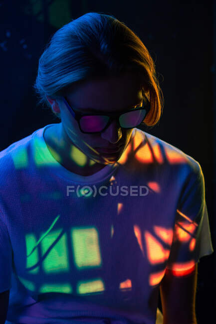 Attractive androgynous guy in stylish sunglasses looking down while sitting under multicolored illumination in dark room — Stock Photo