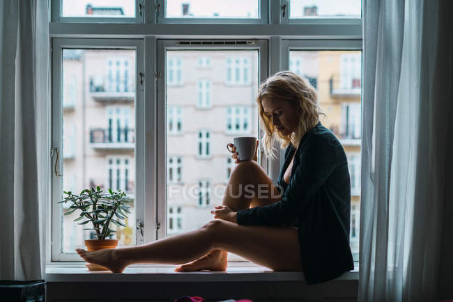 Romântico loira jovem mulher sentada no peitoril da janela com xícara de café — Fotografia de Stock