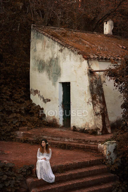 Belle jeune femme afro-américaine en belle robe assise dans la cour près de vieux bungalow terrible dans la forêt et regardant loin — Photo de stock