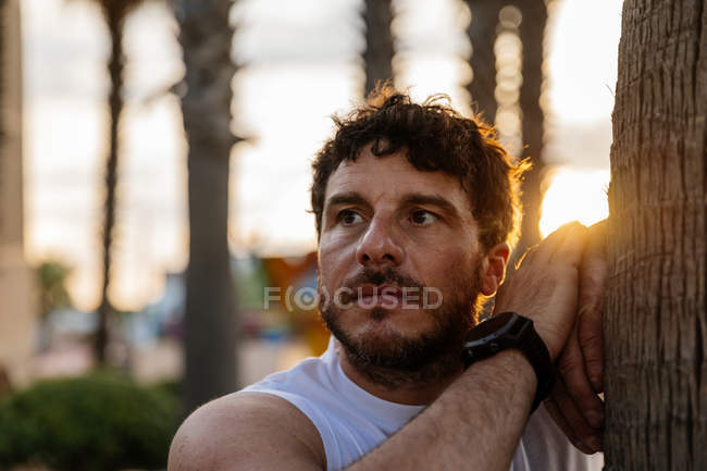 Bearded man in sportswear leaning on tree trunk and looking away during outdoor training in evening — Stock Photo