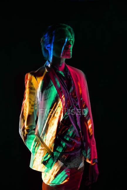 Androgino modello maschile in abito in piedi in posa rilassata sotto illuminazione colorata su sfondo nero — Foto stock