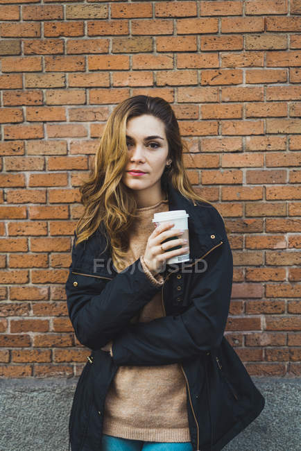 Portrait of young woman with paper cup of coffee standing against brick wall — Stock Photo