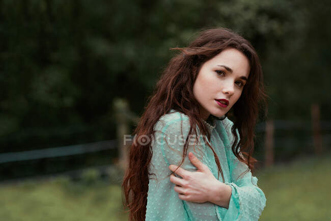 Side view of attractive young lady in elegant dress embracing herself and looking at camera while standing on blurred background of beautiful countryside — Stock Photo