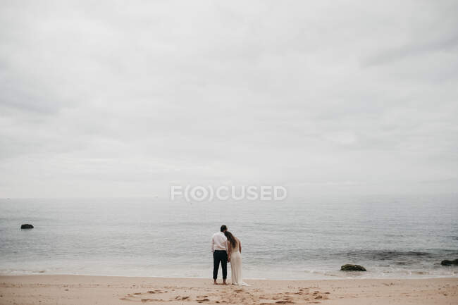 Visão traseira do noivo e da noiva em pé na praia de areia e acariciando contra o céu nublado e paisagem marinha — Fotografia de Stock