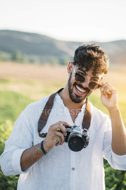 Handsome young guy in sunglasses cheerfully smiling and holding retro photo camera while standing on blurred background of amazing countryside — Stock Photo
