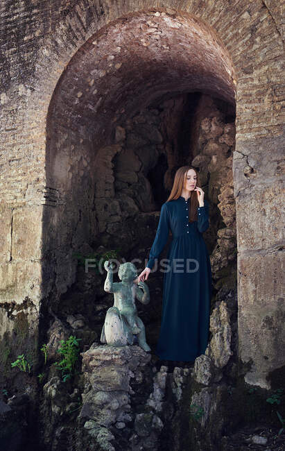 Charming young female in elegant long dress touching small statue and keeping eyes closed while standing in arch of ancient castle — Stock Photo