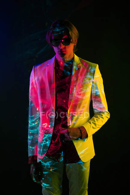 Androgynous male model in suit standing in relaxed pose under colorful illumination on black background — Stock Photo