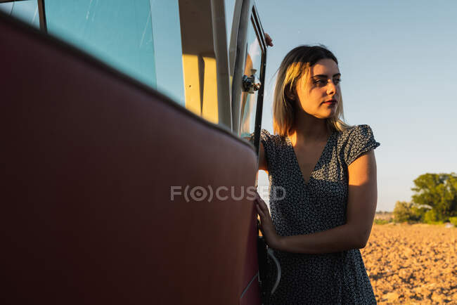 Attraktive junge Frau lehnt an Autotür und blickt in die Kamera, während sie an einem sonnigen Tag in der schönen Natur steht — Stockfoto