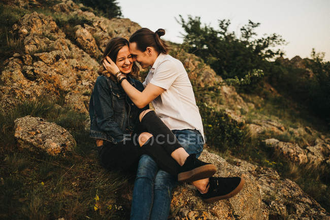 Молодой человек целует девушку в щеку, сидя вместе на склоне скалистой горы — стоковое фото