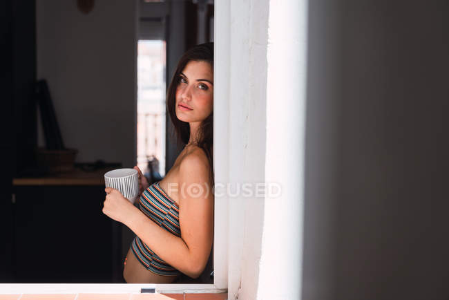 Молода жінка з чашкою кави спирається на стіну біля вікна — стокове фото