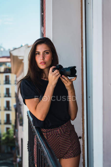 Молода вдумлива жінка стоїть на балконі з фотоапаратом — стокове фото