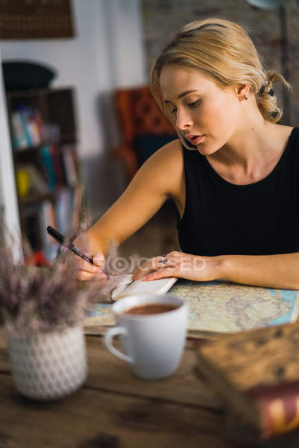 Femme blonde assise à table et planifiant un voyage avec des livres et une carte — Photo de stock