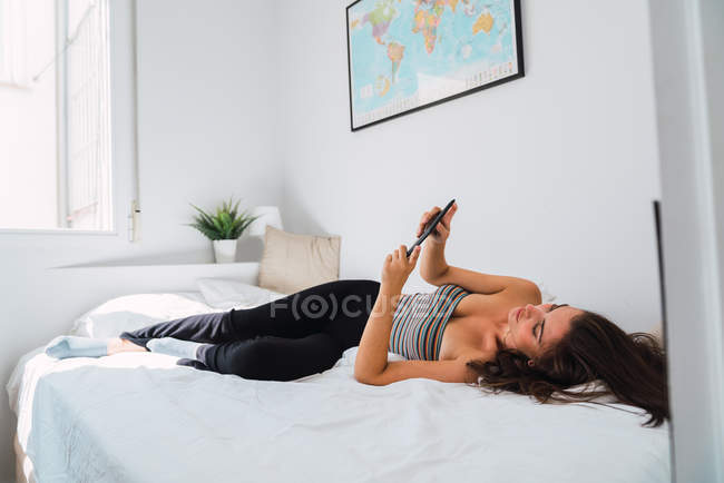 Молода ледача жінка лежить в ліжку і використовує мобільний телефон — стокове фото
