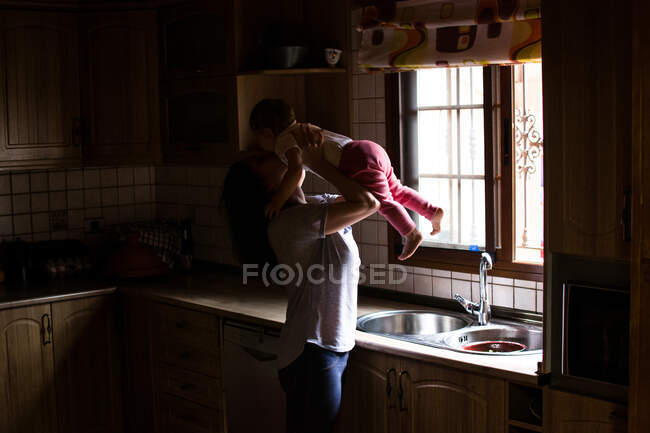 Mujer casual sosteniendo bebé en las manos y besándose con amor de pie en la cocina de la casa - foto de stock