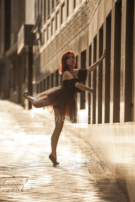 Bailarina de cabeza roja con tutú negro y consejos de ballet rojo bailando en la calle con fondo claro al atardecer - foto de stock