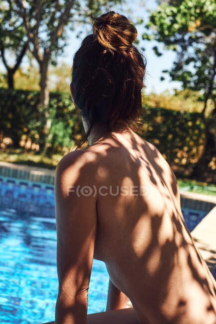 Vista laterale di giovane donna nuda con capelli scuri seduta vicino alla piscina con acqua trasparente blu su sfondo di alberi e cespugli verdi — Foto stock