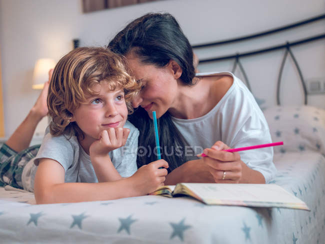 Bella donna e bambino carino sdraiato su un letto confortevole e immagini da colorare nel quaderno insieme — Foto stock
