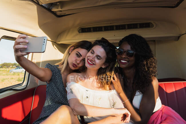 Three cheerful young women sitting inside vintage van and posing for selfie while traveling together — Stock Photo