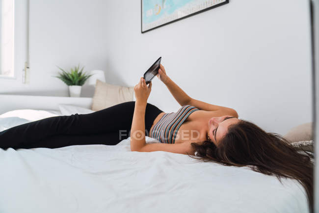 Giovane donna sdraiata a letto e utilizzando il telefono cellulare — Foto stock