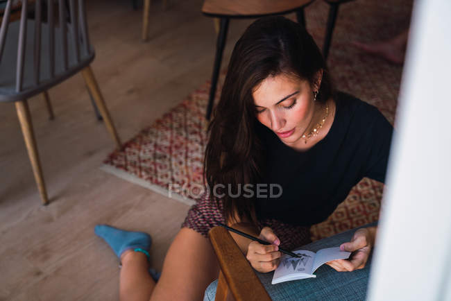 Jovem mulher morena pensativa sentada no chão no quarto e desenhando em pequeno caderno — Fotografia de Stock