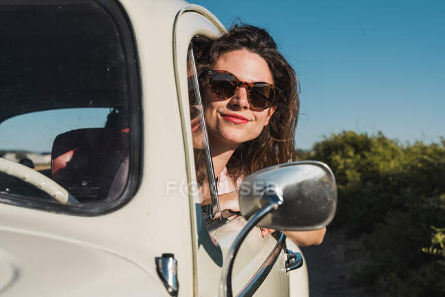 Felice giovane donna in occhiali da sole alla moda guardando fuori dalla finestra dell'auto godendo la luce del sole estiva contro alberi verdi e cielo blu — Foto stock