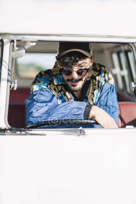 Attractive young man in stylish outfit leaning on steering wheel and looking at camera while sitting inside vintage van during trip in nature — Stock Photo