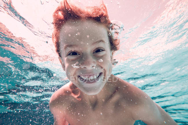 Рыжеволосый ребенок ныряет в воду и смотрит в камеру на фоне прозрачной воды — стоковое фото