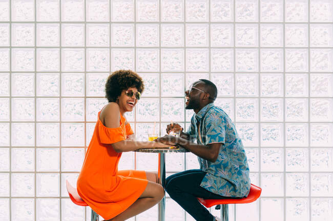 Vista lateral del hombre negro y la mujer sonriendo mientras está sentado en la mesa de la cafetería y tomando algo - foto de stock