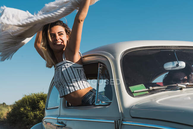 Joven mujer alegre que sobresale de la ventana del coche sosteniendo la bufanda de luz ondulante disfrutando del viaje en la luz del sol brillante - foto de stock