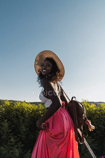 Mujer afroamericana de moda en sombrero llevando maleta y caminando por la carretera rural en verano - foto de stock