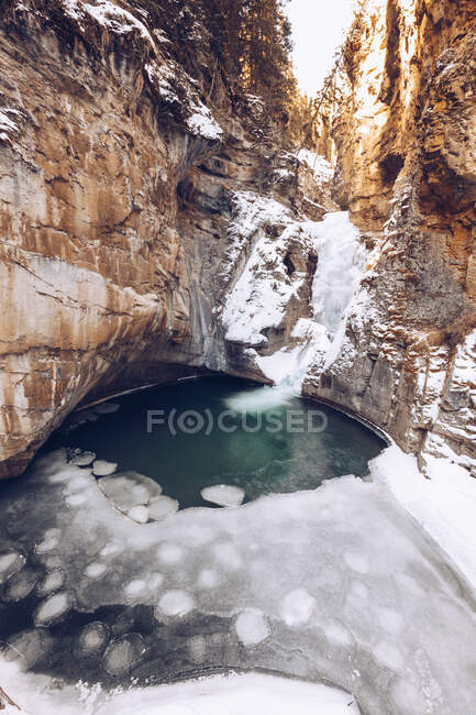River running in melted highland lake at snowy cleft with brown rocks and few winter firs on sunny day — Stock Photo