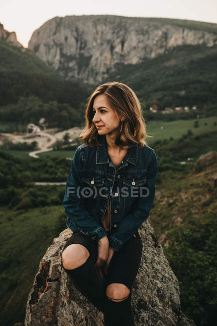 Прекрасна молода жінка посміхається і дивиться геть, сидячи на скелі на фоні мальовничої сільської місцевості — стокове фото