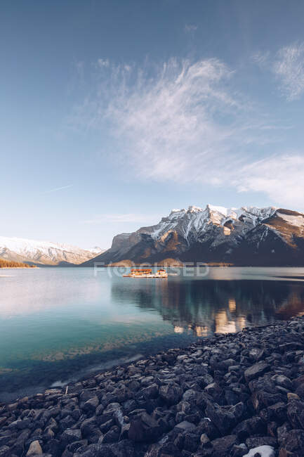 Building with brown roof under sunshine in middle of highland lake with clear blue water on background with picturesque mountains and forest — Stock Photo