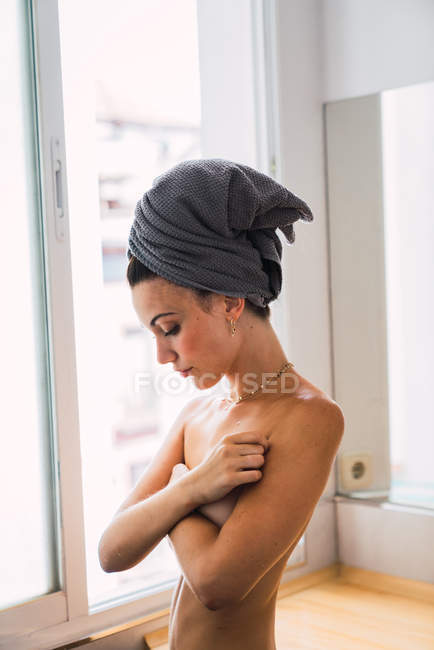 Junge Oben-Ohne-Frau steht mit Handtuch auf dem Kopf im Badezimmer und bedeckt die Brust mit Armen — Stockfoto