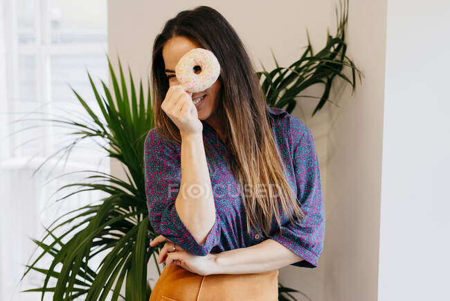 Весела красива жінка стоїть в офісі і дивиться на камеру через дірку пончика . — стокове фото